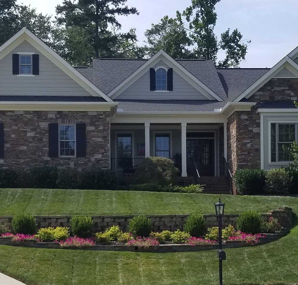 Stone-front home with a mowed lawn, a low retaining wall and a curved bed of pink and yellow flowers